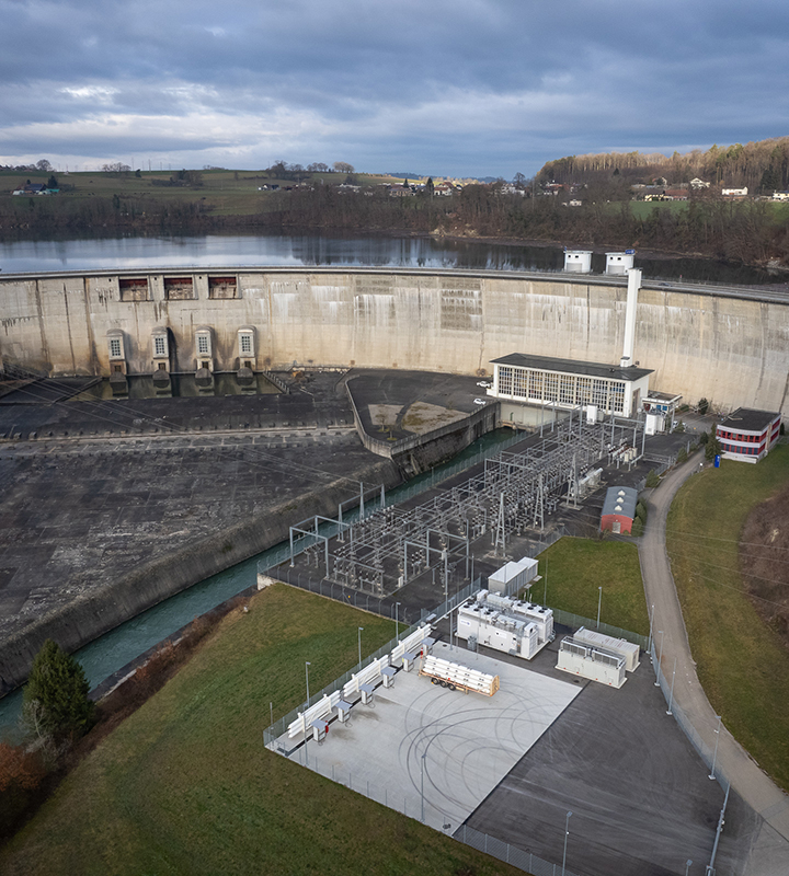Vue aérienne d'un barrage hydroélectrique et de ses installations électriques environnantes, montrant la structure imposante et le bassin d'eau en arrière-plan. Ce site illustre l'infrastructure de production d'énergie renouvelable, un domaine dans lequel Groupe E est impliqué pour fournir une électricité durable et respectueuse de l'environnement.