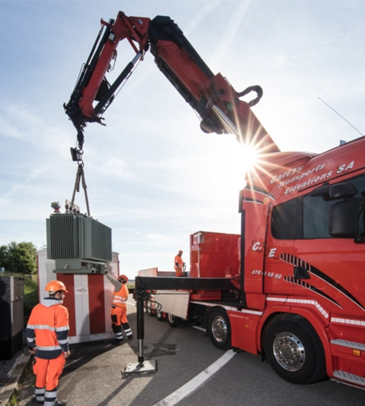 Des collaborateurs de Groupe E supervisent l'installation d'un transformateur électrique à l'aide d'un camion-grue, portant des équipements de sécurité.