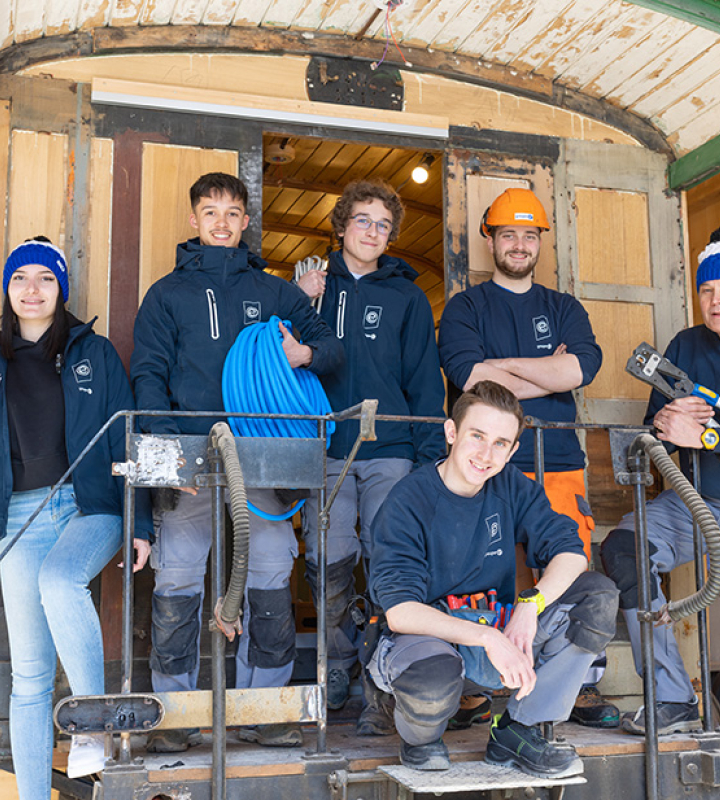 Eine Gruppe von jungen Auszubildenden des Groupe E posiert zusammen auf der Trittstufe einer alten Strassenbahn, die restauriert wird. Alle tragen Arbeitskleidung mit dem Logo des Groupe E, und einige halten Werkzeuge oder elektrisches Material in den Händen. Sie wirken lächelnd und motiviert, was das Engagement des Groupe E für die Berufsausbildung und den Erhalt des Kulturerbes widerspiegelt. Die Holzwerkstatt im Hintergrund verleiht der Arbeitsumgebung eine warme Atmosphäre.