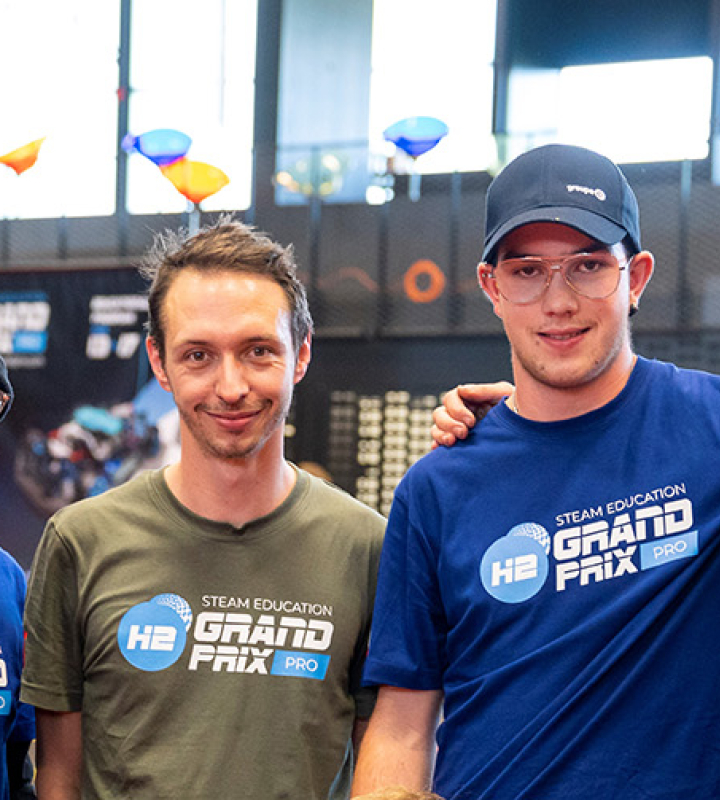 Un groupe de jeunes participants au Grand Prix H2 Steam Education, organisé par le Groupe E, pose ensemble dans un environnement dynamique et convivial. Les participants, habillés de t-shirts et de casquettes avec les logos de l'événement et du Groupe E, montrent leur enthousiasme pour l'initiative. En arrière-plan, d'autres participants et des éléments colorés flottent, illustrant l'énergie et l'innovation promues par le Groupe E dans le domaine de l'éducation et de la technologie.