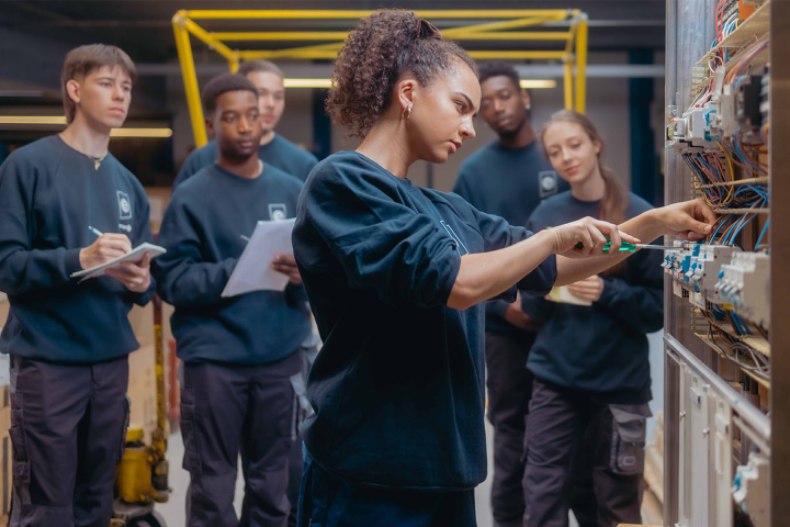 Eine Auszubildende bei Groupe E arbeitet an einem Elektroschaltkasten, aufmerksam beobachtet von ihren Kolleginnen und Kollegen.