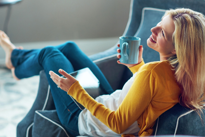 Une femme souriante, détendue dans un fauteuil, tenant une tasse, symbolisant le confort et la sérénité offerts par les solutions énergétiques de Groupe E