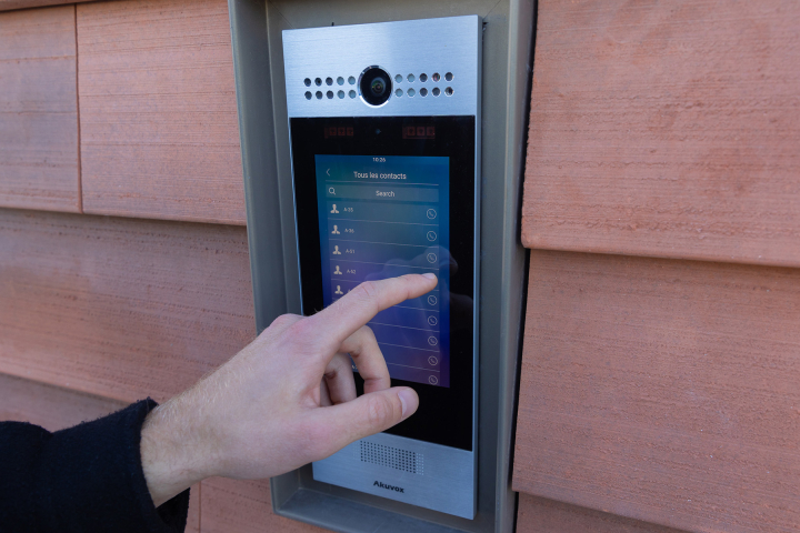 Une main interagit avec une sonnette digitale pour rechercher une personne via l’interface tactile.