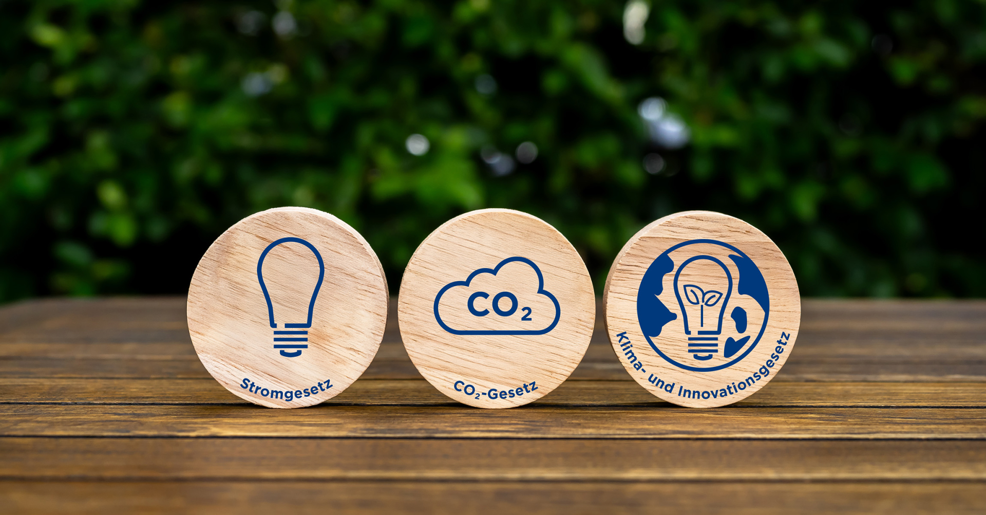 Drei Holzscheiben, die das Stromgesetz, das CO2-Gesetz und das Klima- und Innovationsgesetz mit passenden Symbolen darstellen, auf einem Holztisch.